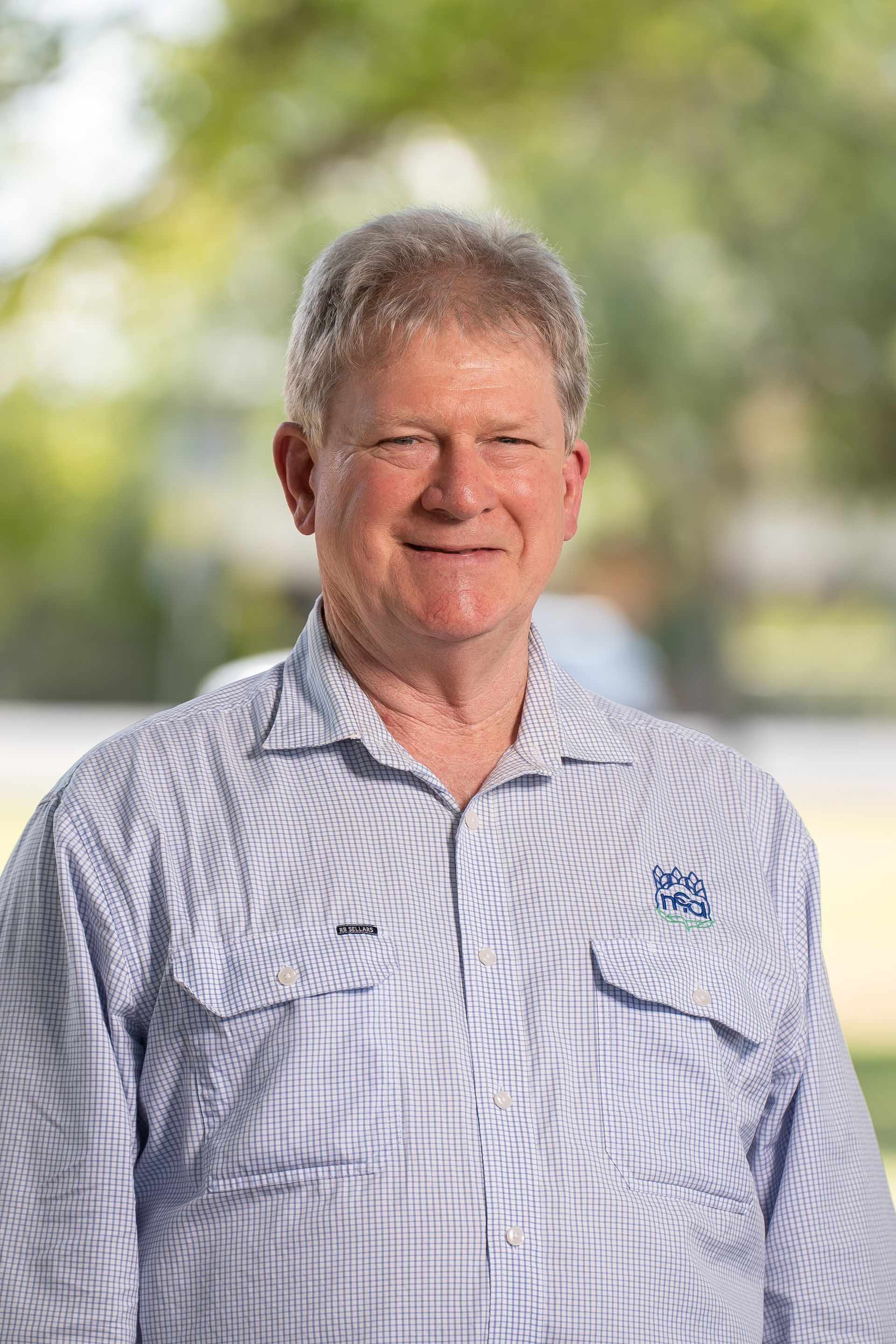 Man in a blue-striped shirt smiles. Outdoors background, appears to be a work uniform.