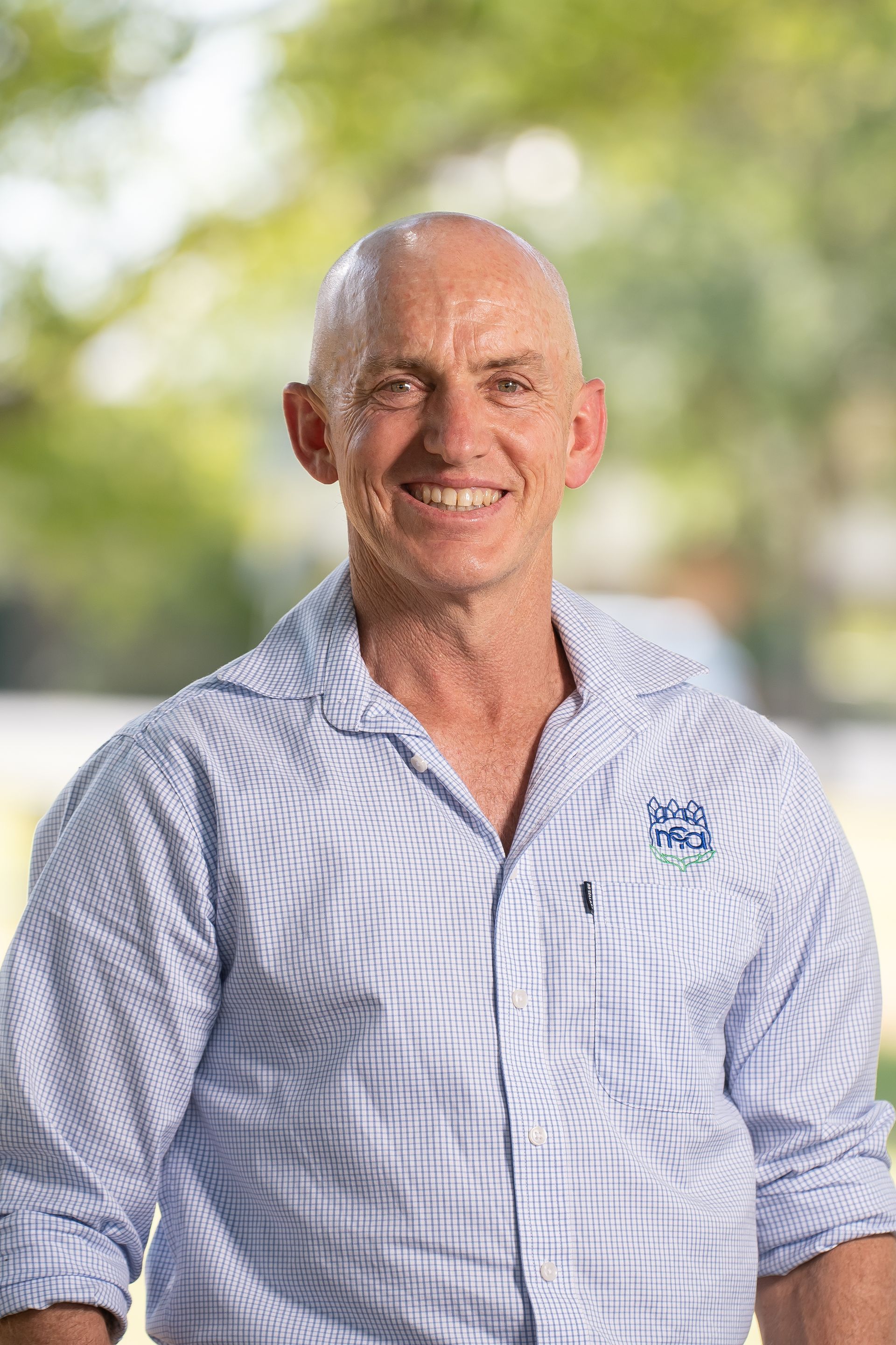 Bald man smiles, wearing a blue button-down shirt with a logo, standing outdoors.