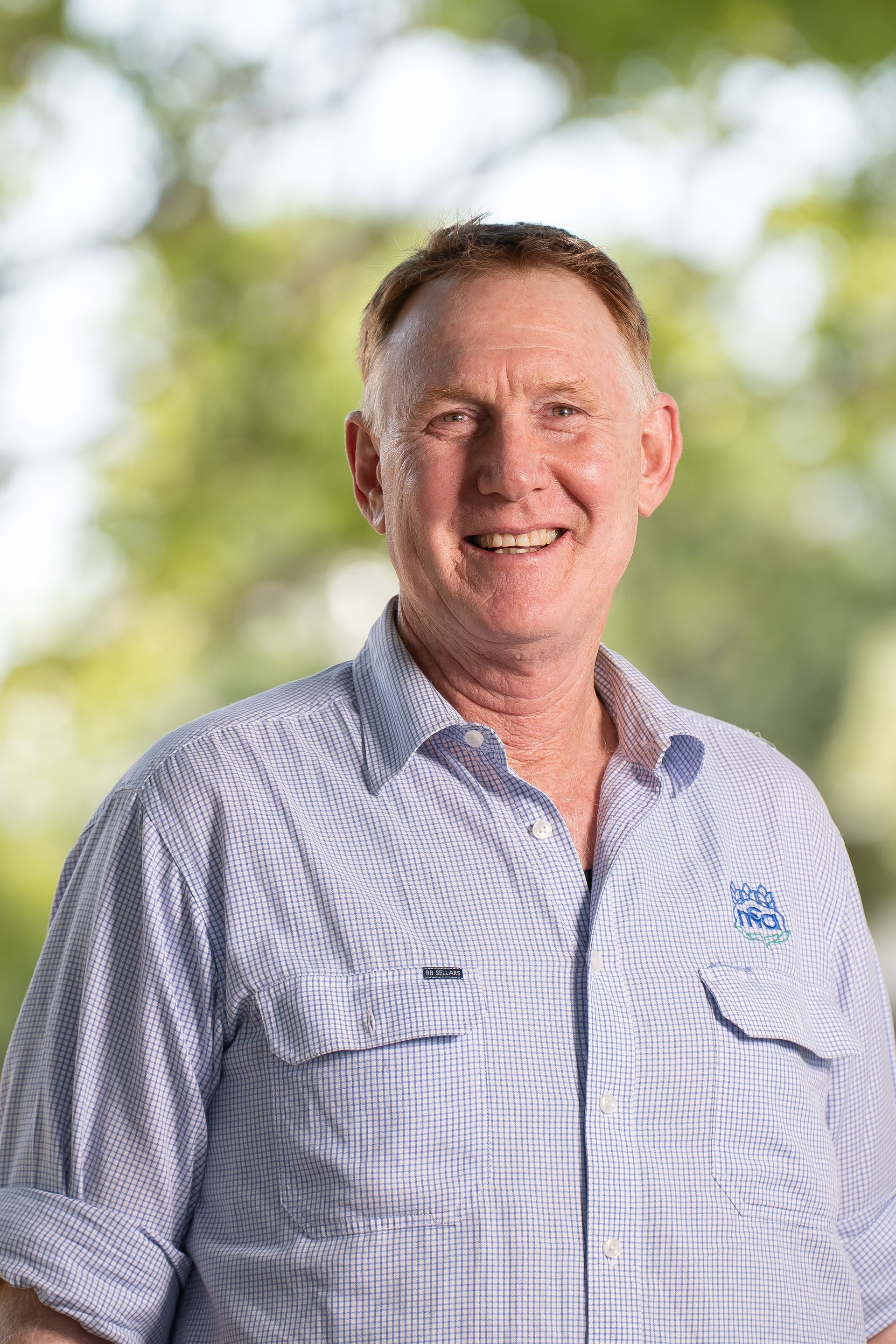Man smiling, wearing a blue checked button-up shirt; trees in the background.