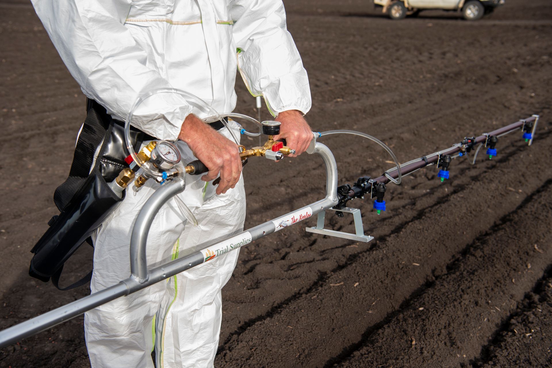 Person in white protective suit spraying a field with a handheld sprayer.