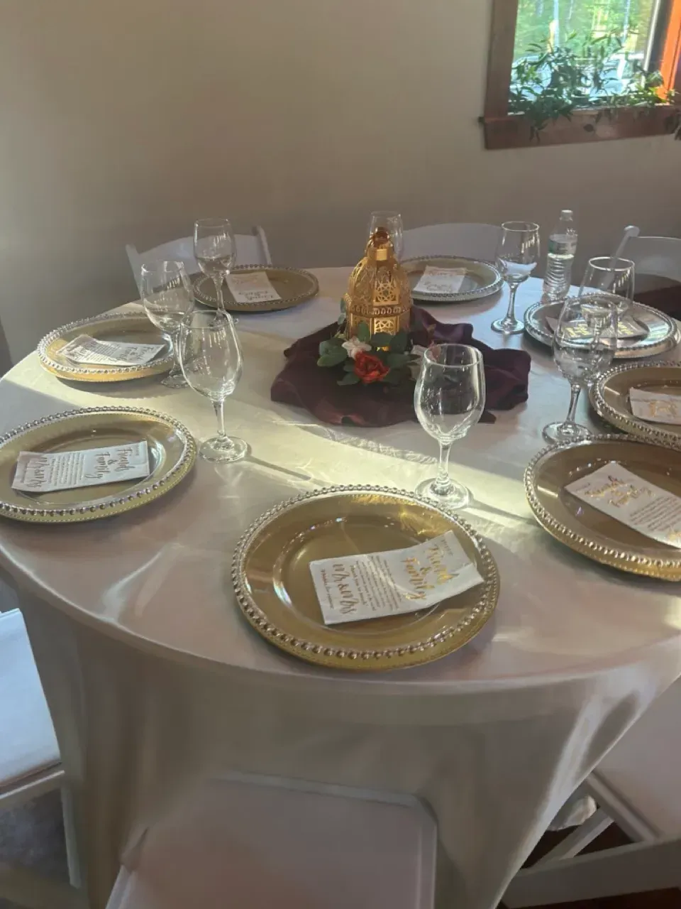 Round dining table set with white tablecloth, gold chargers, wine glasses, and a floral centerpiece.