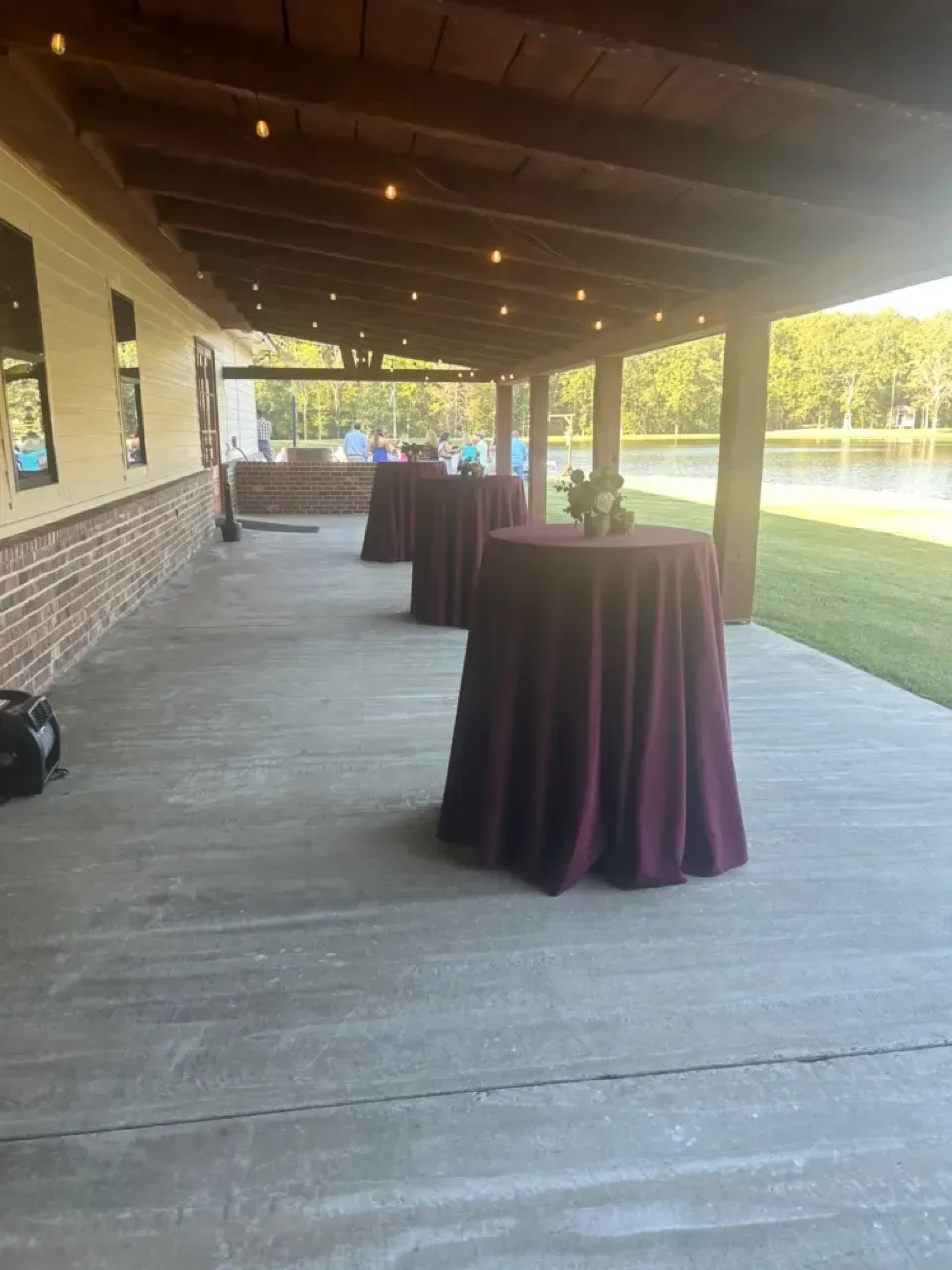 Covered outdoor patio with purple-covered cocktail tables beside a brick building and grassy lawn.