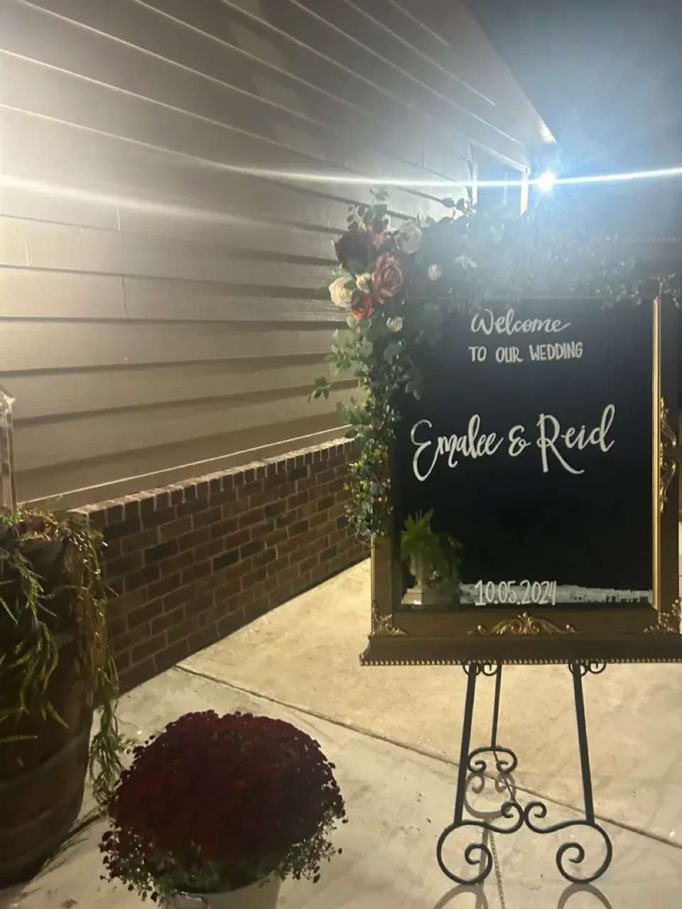 Wedding welcome sign on easel with red roses and greenery beside a brick wall at night