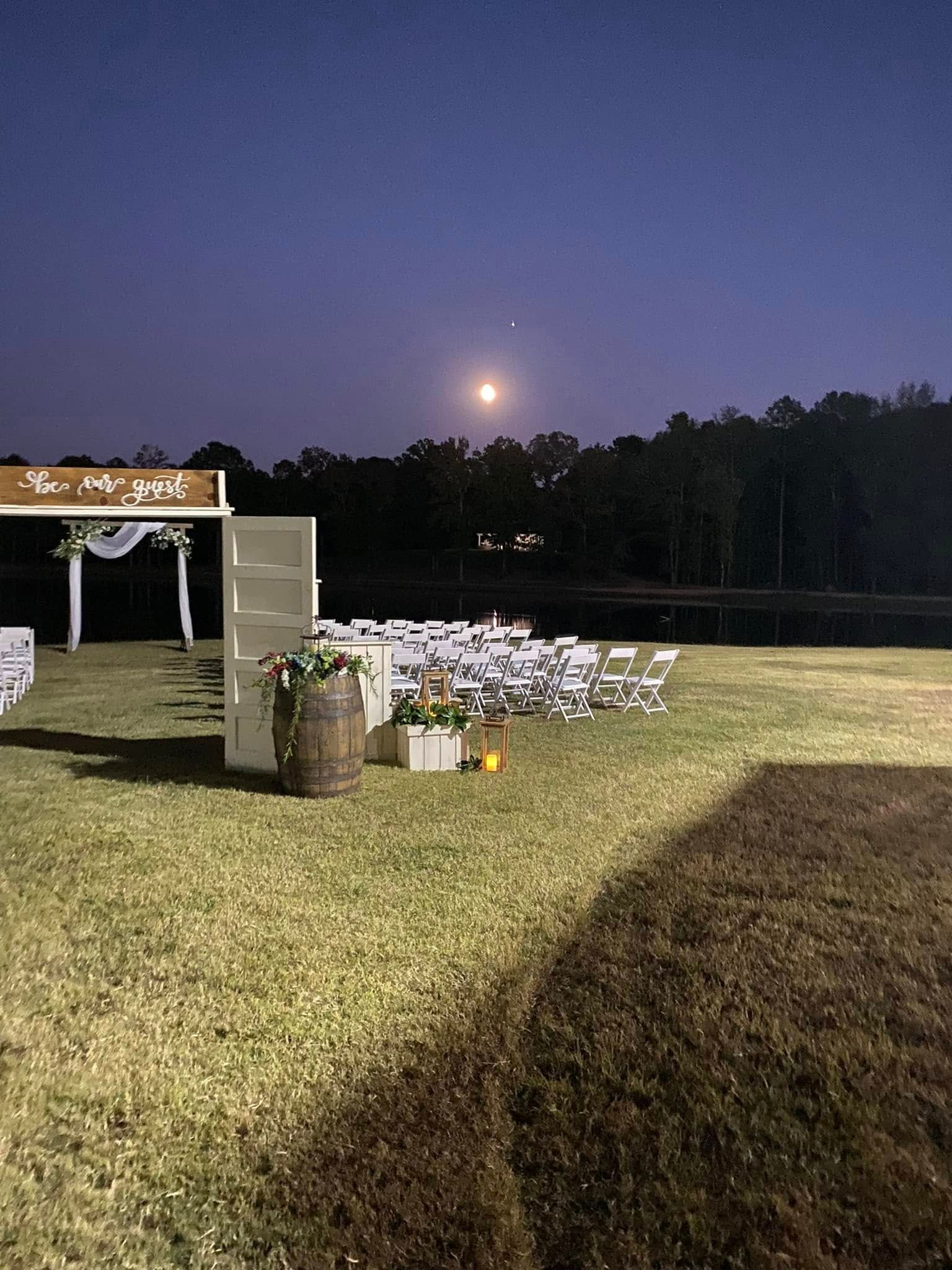 Outdoor wedding setup at dusk with white chairs on a grassy lawn near a lake and gazebo.