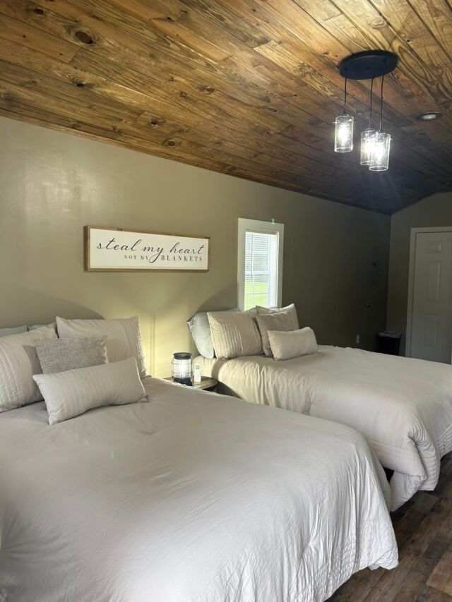 Bedroom with two neatly made beds, wood plank ceiling, window, and wall sign above the headboards