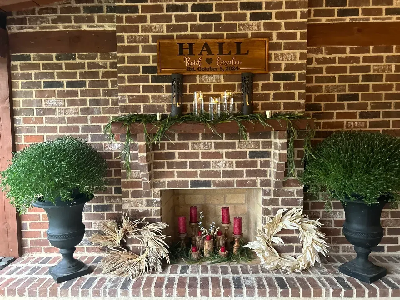 Brick fireplace mantel with “HALL” sign, greenery, candles, and black urn planters.