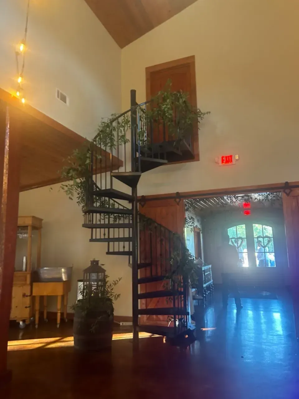 Spiral staircase in a warm-lit hall with wood floors, potted plants, and a bright doorway in the background
