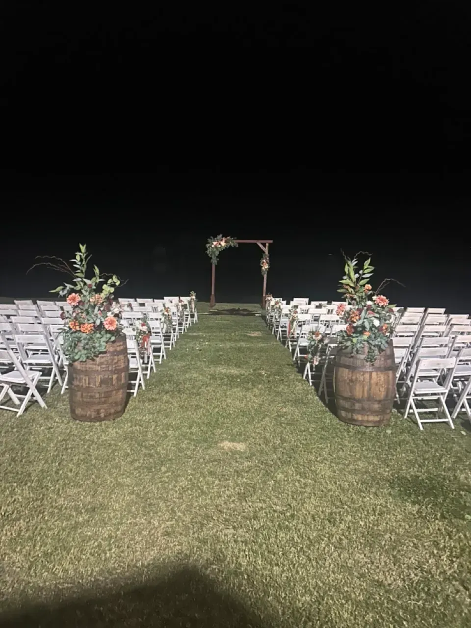 Outdoor wedding aisle with white chairs, barrel flower arrangements, and a floral arch at night