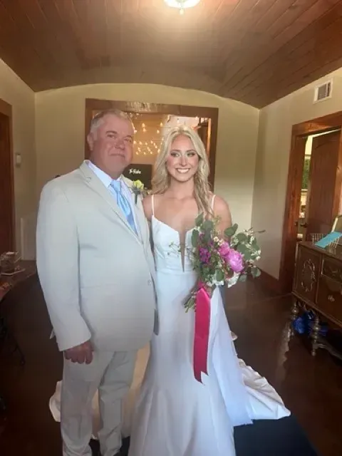 Bride in white dress holding bouquet beside groom in light suit indoors