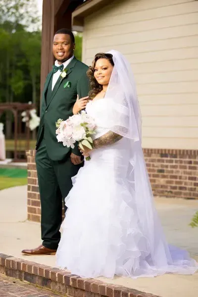 Bride and groom posing outdoors, bride in white gown holding bouquet, groom in dark green suit