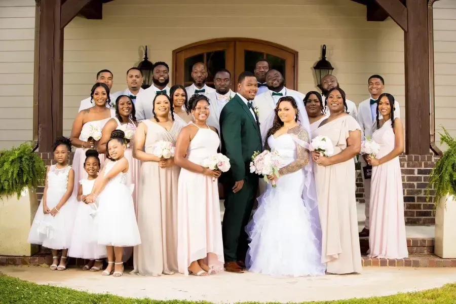 Large wedding party posing on a porch, with bride in a white gown and groom in a green suit.