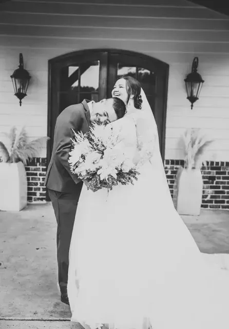 Bride and groom embracing outside a building, with the bride holding a bouquet and wearing a long white gown.