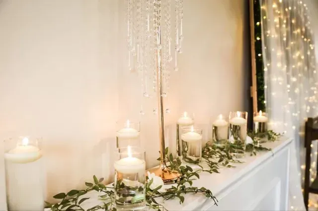 White candles and greenery arranged along a mantel beside sheer fairy lights.