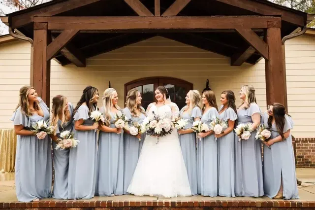 Wedding party posed under a wooden pavilion, bride in white with bridesmaids in gray dresses holding bouquets