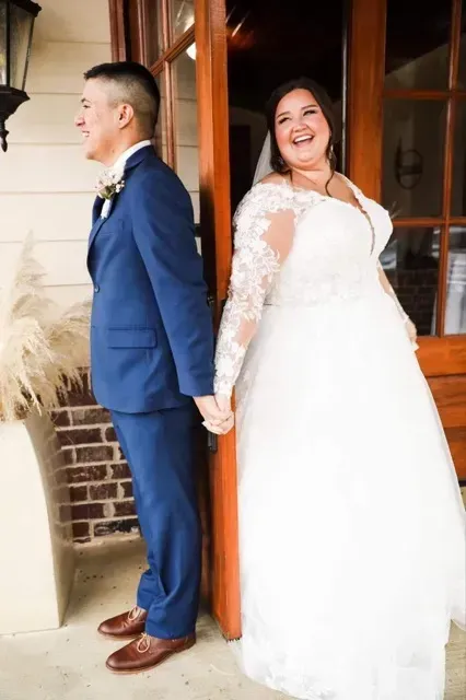 Couple in wedding attire standing back-to-back by a doorway, smiling; bride in white gown, groom in blue suit