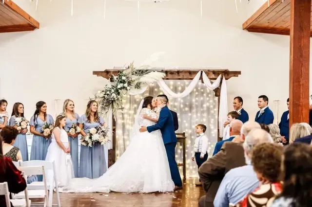 Wedding ceremony with bride and groom kissing under floral arch, attendants and guests watching in a bright hall