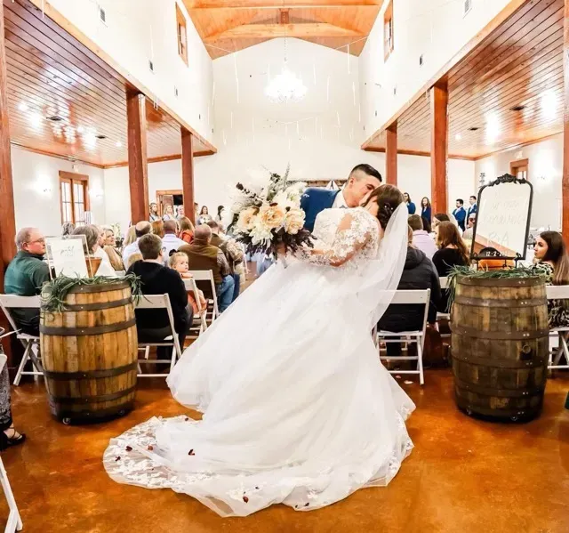 Bride and groom kissing in a rustic barn wedding, with guests seated and floral decor behind them