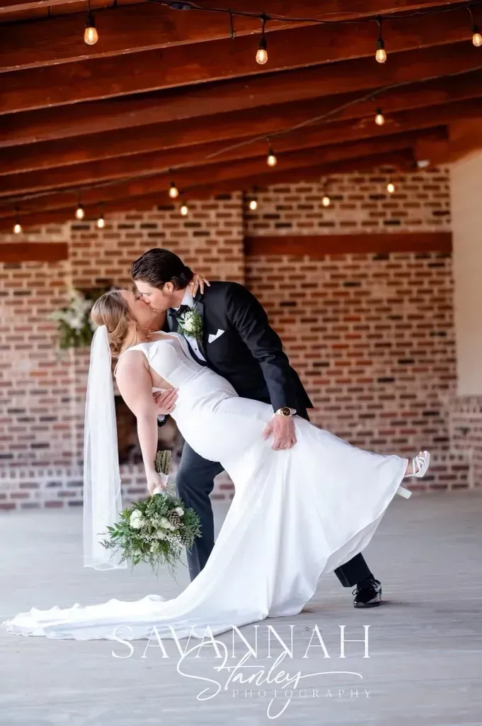 Bride and groom kissing in a white-draped wedding ceremony space with brick walls and string lights