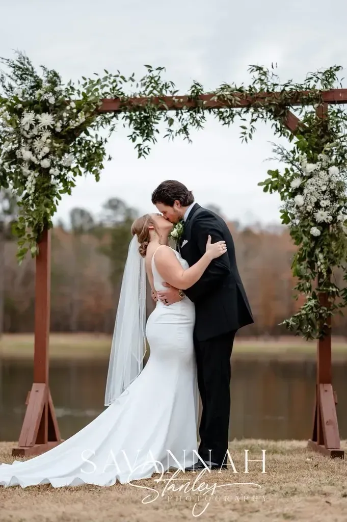 Bride and groom kiss beneath a flower-covered arch by the water.