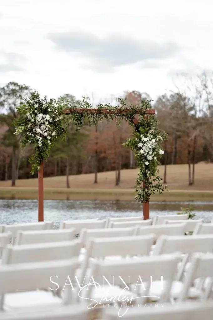 Outdoor wedding ceremony setup with white chairs, floral arch, and pond view