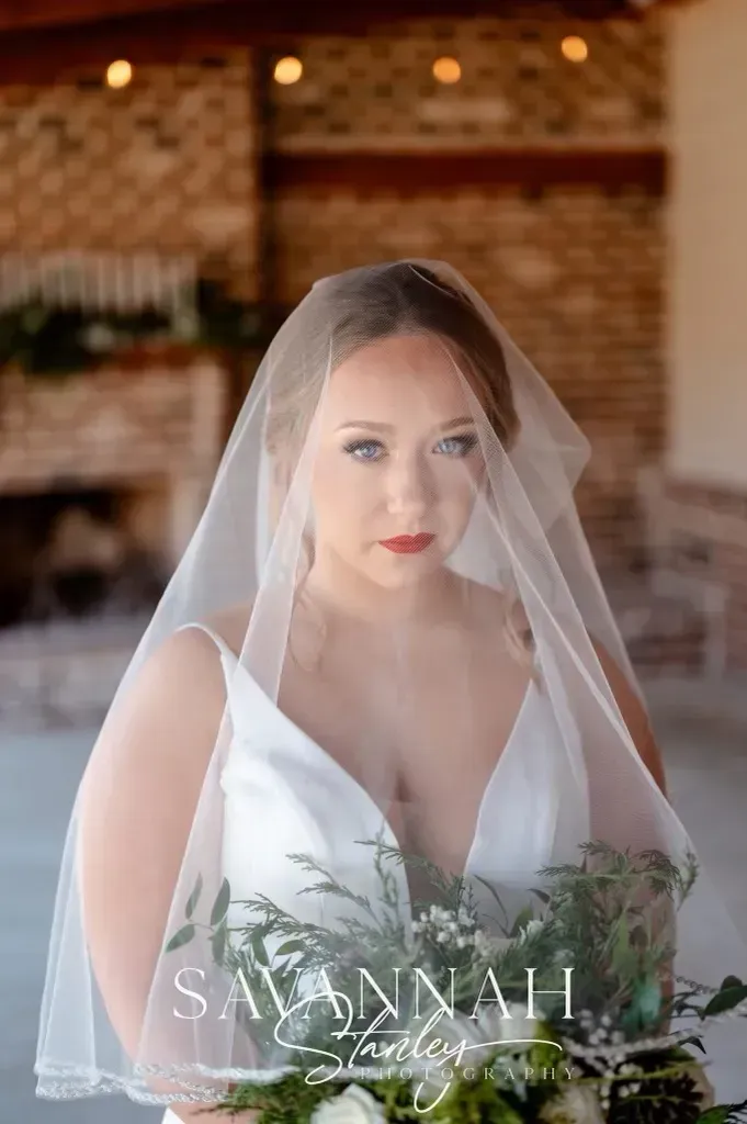 Bride in a white dress and veil holding green foliage, standing indoors with a brick wall background.