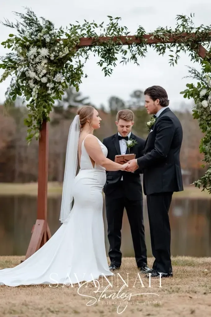Outdoor wedding ceremony under a wooden arch with greenery, bride and groom holding hands, officiant behind them