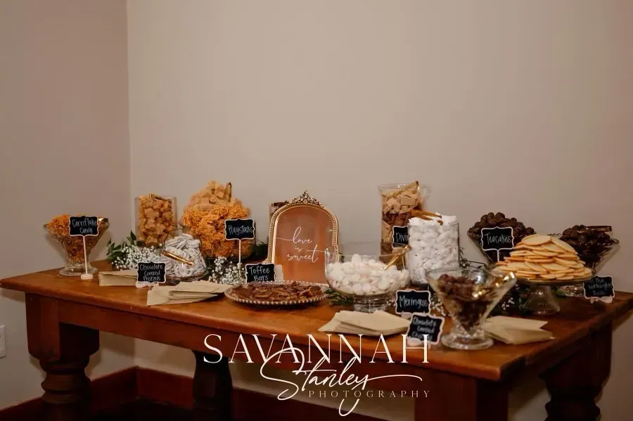 Dessert table with cupcakes, cookies, and a tiered stand on a wooden console table, with “Savannah” sign.