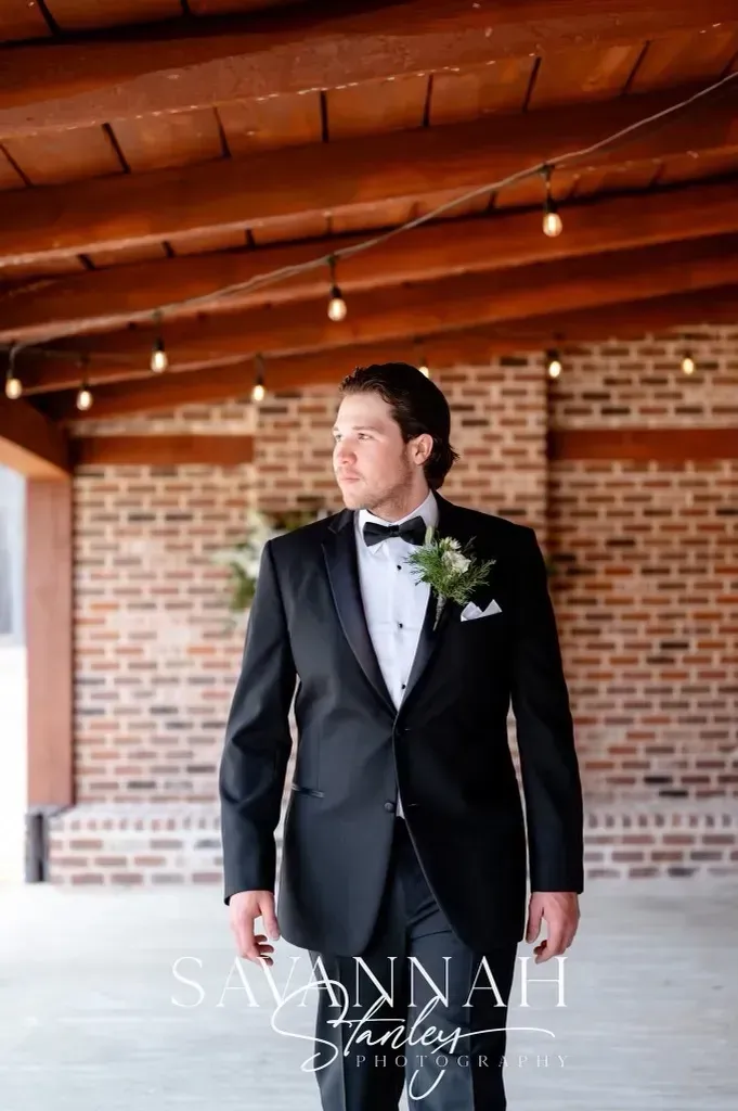 Man in black tuxedo standing under a wooden pavilion with brick walls, looking to the side.