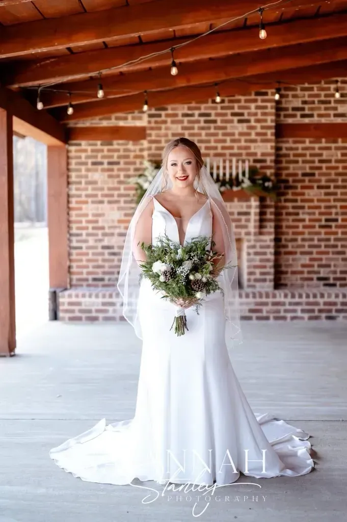 Bride in white gown holding a bouquet, standing in a brick-walled venue with wooden beams