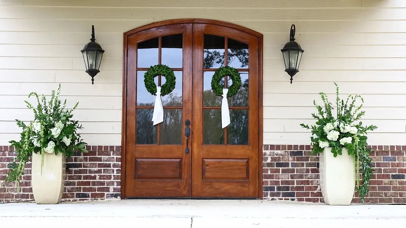 Wooden double front doors with green wreaths, flanked by white urn planters and wall lanterns