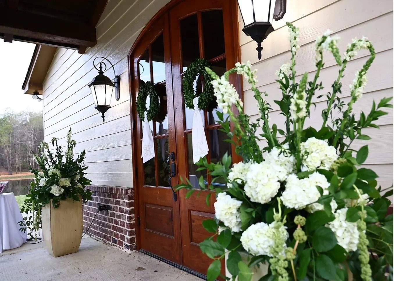 White floral arrangements line a wooden entrance with lanterns and a brick wall outside a building.
