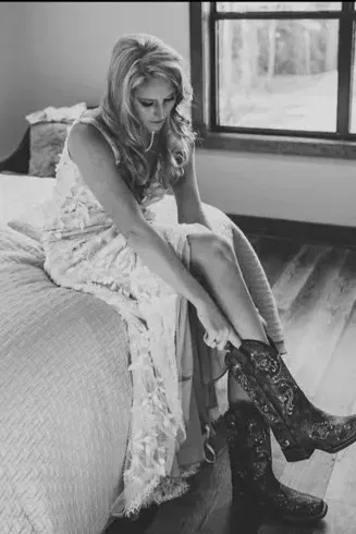 Woman sitting on a bed, pulling on black lace-up boots in a bright bedroom.
