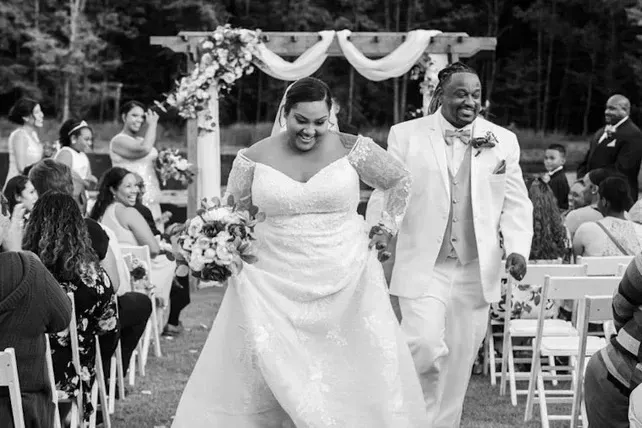 Newlyweds walking down the aisle outdoors, smiling beneath a floral arch at a wedding ceremony.