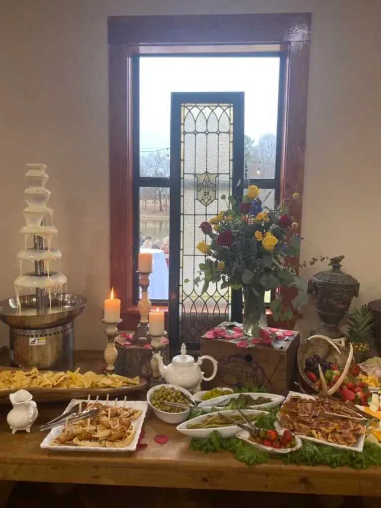 Table set with food, candles, flowers, and a chocolate fountain in a bright room by a window.