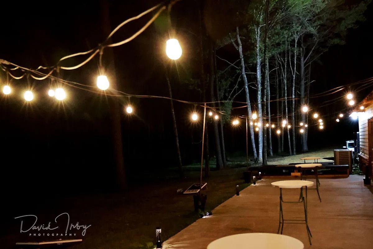 String lights over a wooden patio at night with trees and a few tables in the background
