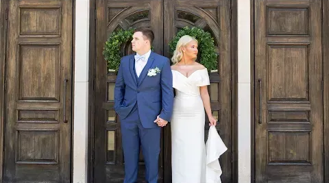 Bride and groom standing in a doorway outside a wooden building, holding hands