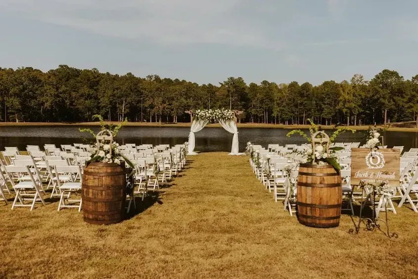 Outdoor wedding ceremony setup with white chairs lining a grassy aisle and a floral arch near a lake