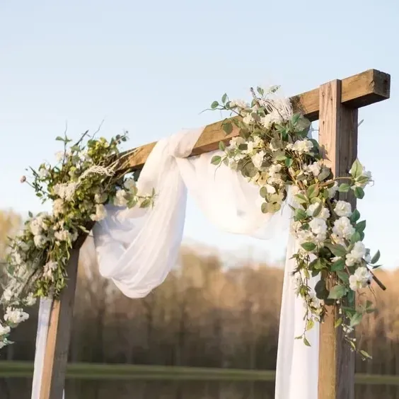 Floral-draped wooden wedding arch with white fabric outdoors at sunset