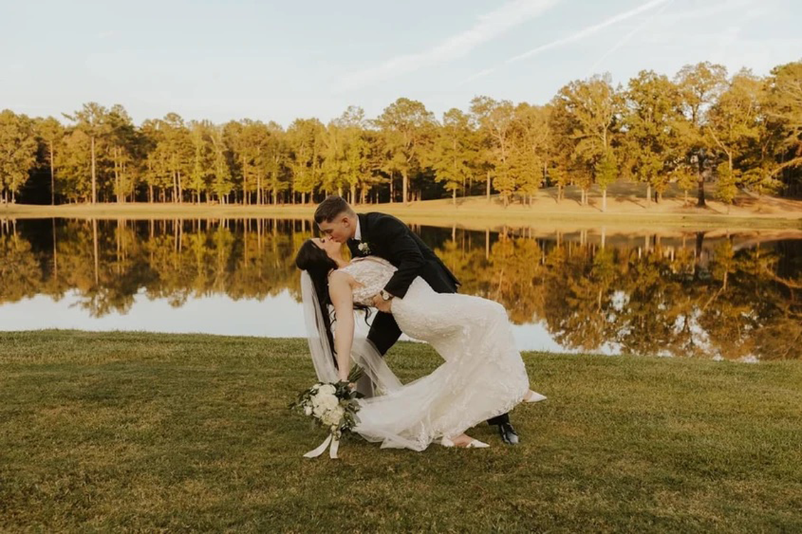Bride and groom kiss on a grassy lawn by a calm lake at golden hour