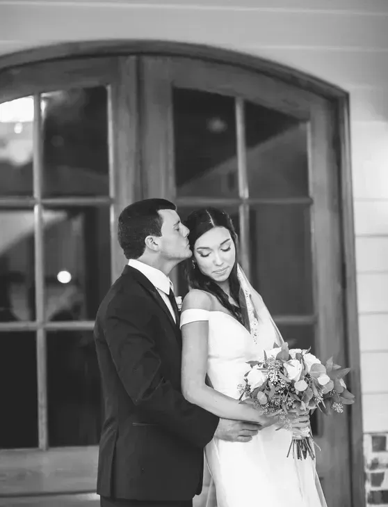 Couple in wedding attire posing by an arched window, bride holding a bouquet.