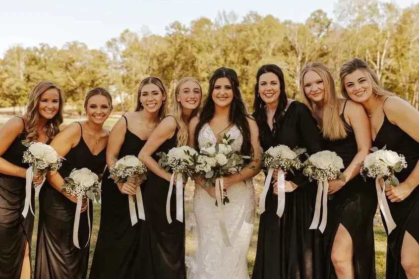 Bridal party in black dresses and a white gown holding bouquets outdoors in autumn light