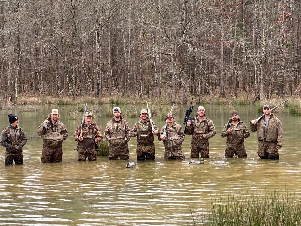 Nine people in camouflage stand waist-deep in muddy water near a wooded bank, posing with rifles.