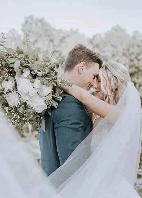 Bride and groom embracing outdoors, with bouquet and flowing veil in a soft white floral setting
