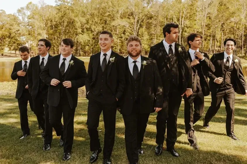 Group of people in black suits walking together outdoors on a grassy field with trees behind them