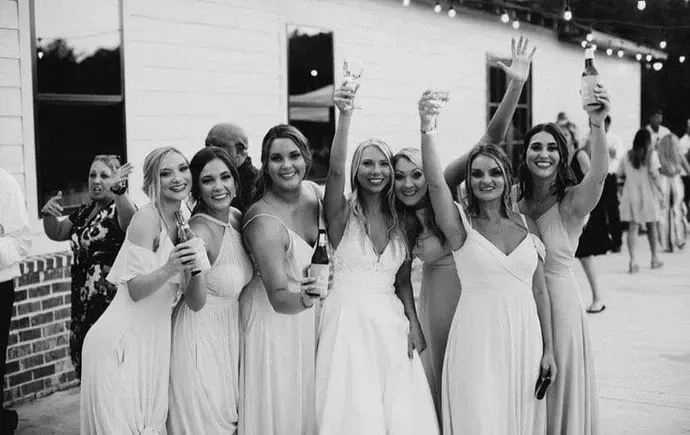Women in white dresses posing outdoors, smiling and holding drinks at a celebration