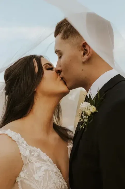 Bride and groom kissing outdoors under a white veil, with the groom in a black suit and boutonniere