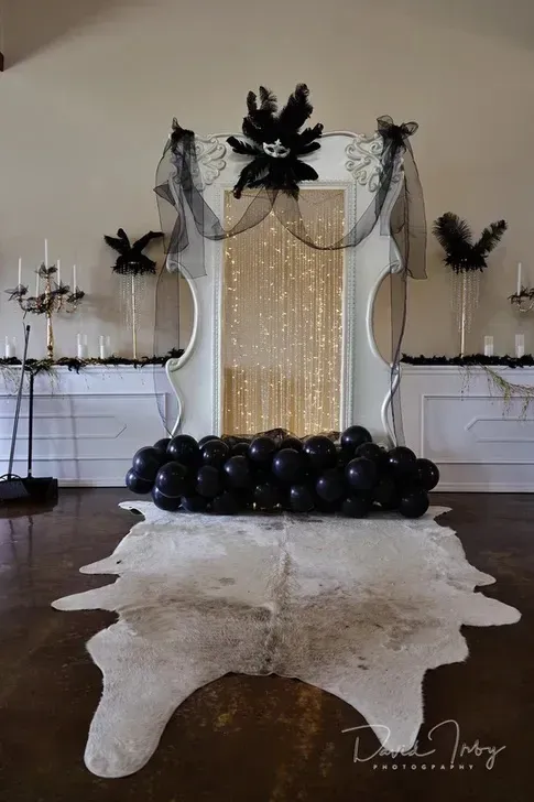 Black-and-white party backdrop with balloons, draped fabric, candles, and a white cowhide rug on a wooden floor