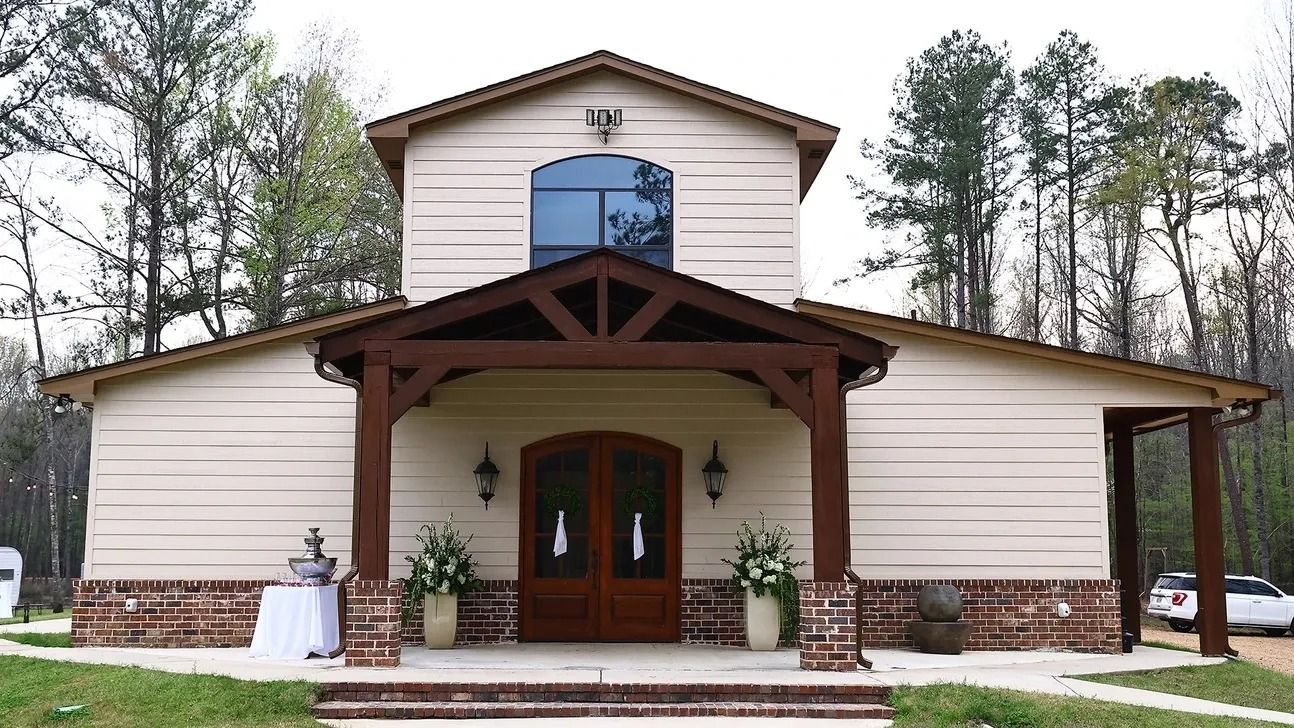 Front view of a beige church with a brown roof, arched wooden doors, and stone trim, surrounded by trees.