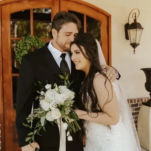 Bride and groom embracing at a doorway, holding a white bouquet, both smiling.