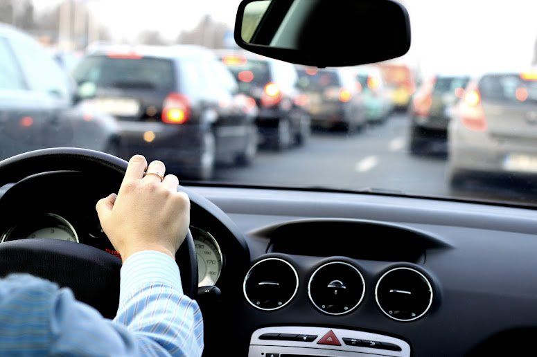 Hand on steering wheel, driving in heavy traffic. Cars lined up on highway, gray, and black vehicles.